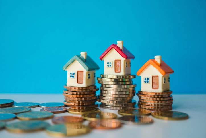Three small toy houses resting on top of stacks of coins against a solid blue background
